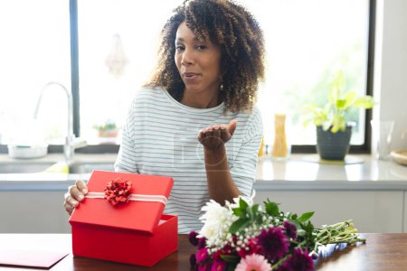 Biracial woman presents a gift at home during an online date video call. She's seated at a table with flowers, expressing joy and anticipation.