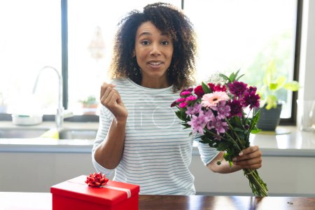Biracial woman holding flowers in a bright kitchen during an online date video call. She seems pleasantly surprised by a thoughtful gift on the counter.