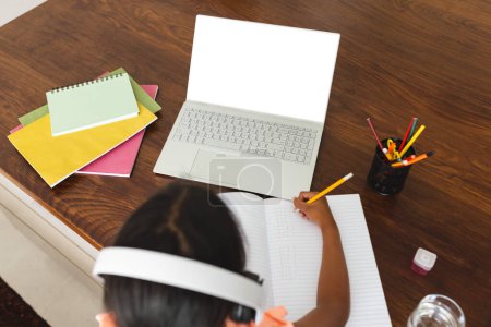 Biracial girl studying at home using a laptop, with copy space. She's focused on her notebook, surrounded by school supplies.