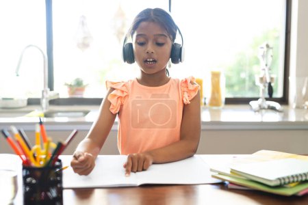 Biracial girl studies at home on a video call, engaged in online learning. She's focused on her homework with headphones on.