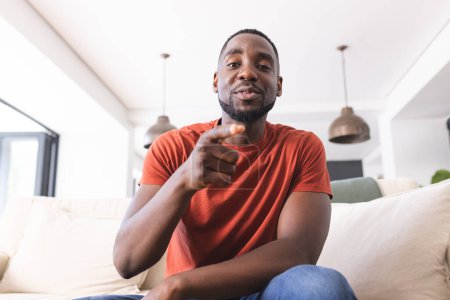 African American man in a red shirt is holding a remote and relaxing on a couch on video call. He appears comfortable and engaged, watching television in a well-lit living room.