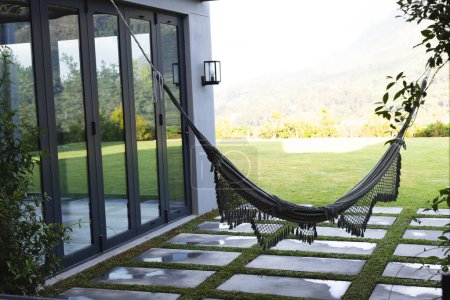 Una hamaca negra cuelga invitadamente en el patio trasero de una casa moderna con espacio de copia. Exuberante vegetación y montañas en la distancia crean un entorno al aire libre sereno.