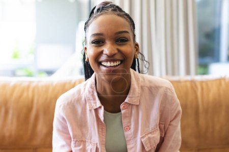 A young African American woman smiles brightly, seated on a tan sofa on video call. Her joyful expression and casual attire create a relaxed, welcoming atmosphere.