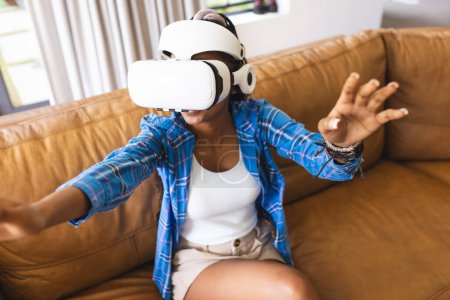 Young African American woman explores virtual reality, gesturing with her hands. She's wearing a VR headset in a bright living room, immersed in a digital experience.