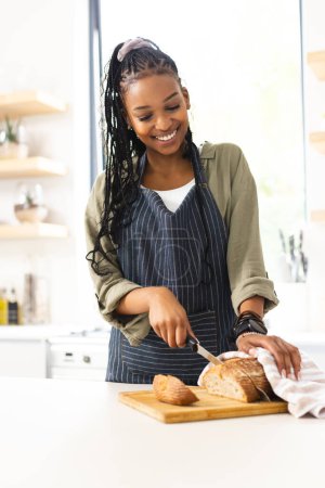 A young African American woman slices bread in a bright kitchen. She wears a striped apron over casual clothes, her braided hair styled neatly, radiating joy in her domestic setting.