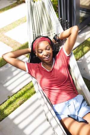 Eine junge Afroamerikanerin entspannt sich in einer Hängematte und genießt Musik. Sie trägt ein rotes T-Shirt, blaue Shorts und lächelt zufrieden in einer sonnigen Outdoor-Umgebung.