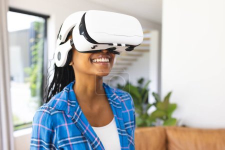 Young African American woman wears a virtual reality headset, smiling in a bright room. Dressed in a blue plaid shirt, she explores a digital world, experiencing modern technology at home.