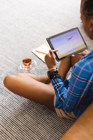 Young African American woman is using tablet while sitting on floor to book vacation online. She's dressed casually in blue plaid shirt, with notebook and drink beside her.