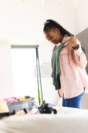 Young African American woman packs a suitcase, deciding on clothes with copy space. She is in a bedroom, preparing for a trip with a camera on the bed indicating a vacation.