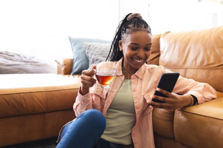 Young African American woman enjoys tea while browsing her phone on a couch. She's wearing a casual pink shirt and jeans, exuding a relaxed vibe in a cozy home setting.