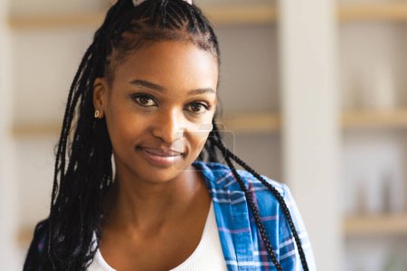 Eine junge Afroamerikanerin mit geflochtenem Haar lächelt sanft in die Kamera. Sie trägt ein weißes Oberteil mit einem blau karierten Hemd, das ein entspanntes und nahbares Auftreten vermittelt.