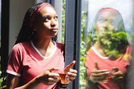 Eine junge Afroamerikanerin blickt aus einem Fenster und hält eine Tasse Tee in der Hand. Ihr reflektierender Ausdruck und das natürliche Licht schaffen eine heitere, besinnliche Atmosphäre.