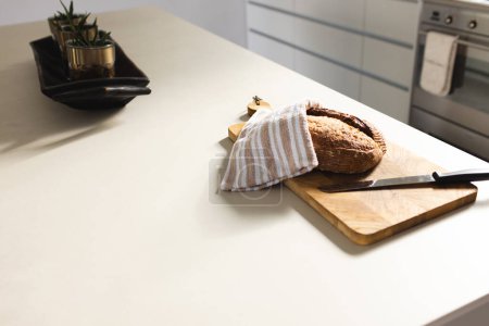A loaf of bread is partially covered with a striped cloth on a kitchen counter with copy space. Nearby, a wooden cutting board and knife suggest the bread is ready to be sliced and served.