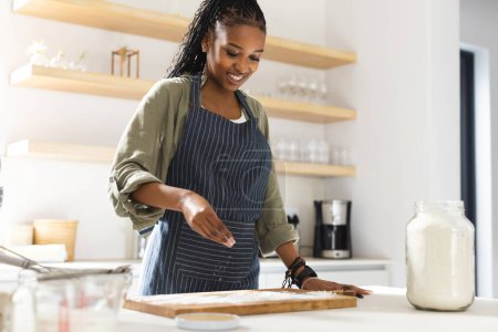 A young African American woman seasons food in a bright kitchen. She wears a striped apron over casual clothes, her hair styled in braids, exuding a joyful cooking experience.