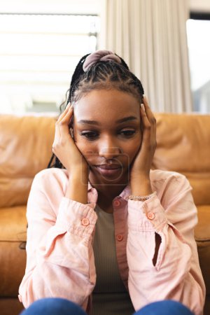 A young African American woman feeling sad, resting her face in her hands. She's seated indoors, her expression conveying a sense of contemplation or concern.
