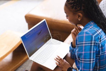 Una joven afroamericana se centra en la pantalla de su computadora portátil, reservando unas vacaciones en línea. Está adentro, con una camisa a cuadros azul, con el pelo peinado en trenzas..