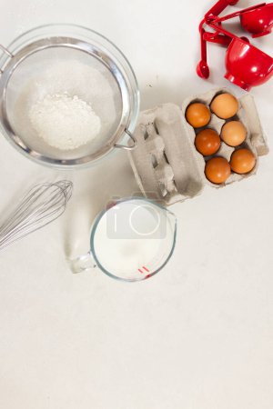 Ingredients for baking are neatly arranged on a white surface with copy space. A carton of eggs, a jug of milk, a sifter with flour, and measuring cups set the stage for a baking session.
