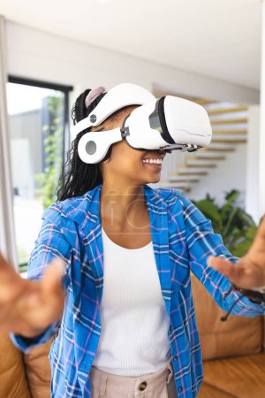 Young African American woman explores virtual reality, wearing a VR headset. She is smiling, dressed in a blue plaid shirt and reaching out with her hands, immersed in the experience.