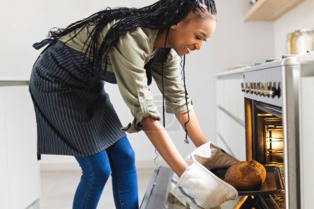 A young African American woman takes a loaf of bread out of an oven. She is wearing oven mitts, a striped apron, and a joyful expression, suggesting a successful baking endeavor at home.