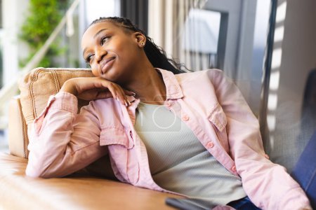 A young African American woman lounges thoughtfully on a sofa. She wears a pink jacket over a gray top, her hair styled in long braids, exuding a relaxed and pensive mood.