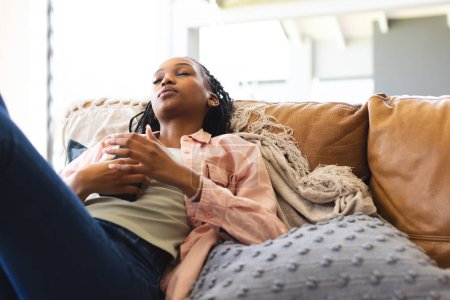 A young African American woman relaxes on a couch, eyes closed and hands clasped. She wears a pink shirt and jeans, exuding a sense of calm in a cozy home setting.