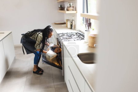 A young African American woman checks on food in an oven with copy space. Dressed casually, she is cooking in a modern kitchen setting, showcasing everyday domestic life.
