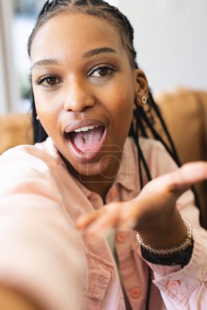 A young African American woman with braided hair is taking a selfie. She wears a pink shirt and exhibits a playful expression in a brightly lit indoor setting.