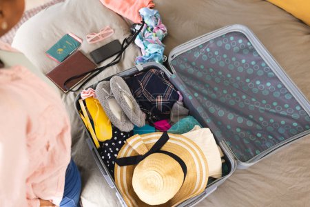 A young African American woman is packing a suitcase with various items. Clothing, a passport, and a sun hat indicate preparation for a trip, a vacation.