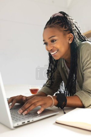 Una joven afroamericana sonríe mientras usa un portátil. Ella está vestida casualmente, su cabello trenzado y su reloj de pulsera complementando su comportamiento alegre en el trabajo..