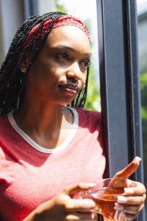 A young African American woman gazes out a window, holding a cup of tea. Sunlight filters through, highlighting her braided hair with red accents and thoughtful expression.