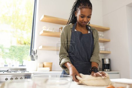 A young African American woman kneads dough on a kitchen counter with copy space. She is focused on her task, wearing a striped apron in a well-lit, modern kitchen setting.