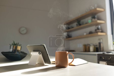 Dampf steigt aus einem Kaffeebecher neben einem Tablet auf einer Küchentheke mit Kopierraum. Warmes Sonnenlicht filtert durch, was auf eine friedliche Morgenroutine zu Hause hindeutet.