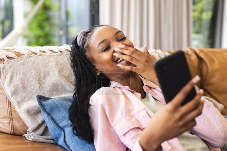 Young African American woman laughs while looking at her phone, reclining on a couch. Her joy is palpable as she enjoys a casual moment in a cozy indoor setting.