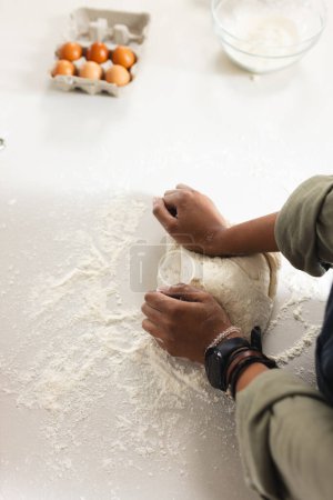 A young African American woman kneads dough on a floured surface with copy space. Eggs and a bowl of flour suggest baking preparations in a home kitchen setting.