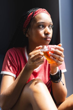 A young African American woman enjoys a cup of tea, gazing thoughtfully. She sports a red headband, a casual t-shirt, and a contemplative expression in a serene indoor setting.