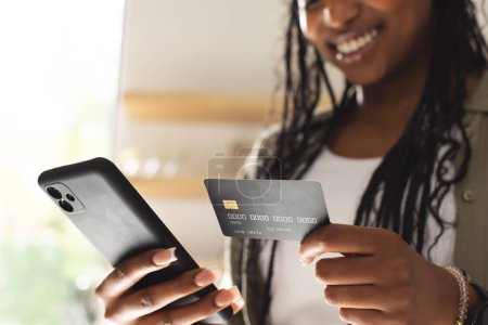 Eine junge Afroamerikanerin lächelt, während sie eine Kreditkarte und ein Smartphone in der Hand hält. Sie scheint einen Online-Kauf zu tätigen, der ein Gefühl von modernem Konsum und Bequemlichkeit ausstrahlt..