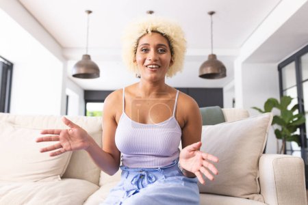 Young biracial woman with curly blonde hair sits on couch, gesturing while talking on a video call. She wears a light blue tank top and denim shorts, creating a casual atmosphere in a bright living room.