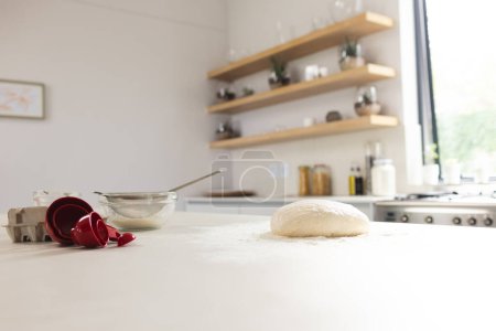 A kitchen counter is dusted with flour, featuring dough and a red measuring cup with copy space. The scene suggests baking preparation, with ingredients and utensils neatly arranged on the surface.