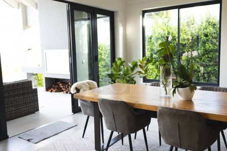 A modern dining room features a large wooden table with grey chairs and a fireplace, with copy space. Sunlight filters through the windows, highlighting the indoor plants and creating a cozy atmosphere.
