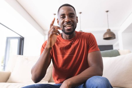 Afroamerikaner im roten Hemd gestikuliert mit dem Zeigefinger und lächelt bei einem Videoanruf herzlich. Indoor-Ambiente mit gemütlichem Ambiente suggeriert ein ungezwungenes, freundliches Gespräch oder Ideenaustausch.