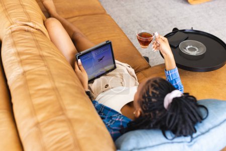 Young African American woman lounges on sofa with tablet and drink, booking vacation online. She is relaxing at home, enjoying her leisure time in a casual setting.