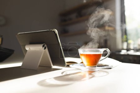Steam rises from a hot cup of tea next to a tablet on a sunlit table. The scene suggests a peaceful morning with a moment for relaxation or catching up on news.