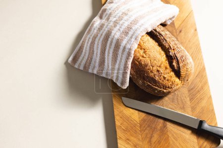 A freshly baked loaf of bread rests on a wooden cutting board beside a knife, with copy space. The bread is partially covered with a striped cloth, evoking a sense of home-cooked warmth.