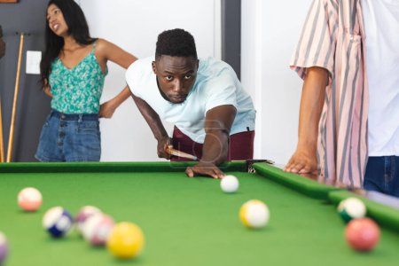 Young African American man focuses on a pool shot, with a young biracial woman watching. Casual indoor setting, the group enjoys a game of billiards, showcasing leisure time.