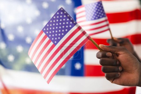 Mano afroamericana sosteniendo una bandera americana con otra bandera en el fondo. El enfoque en la bandera simboliza el patriotismo y el orgullo nacional.