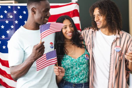 African American man and biracial couple hold American flags, all wearing I Voted stickers. They stand before a large American flag, smiling, having participated in an election.