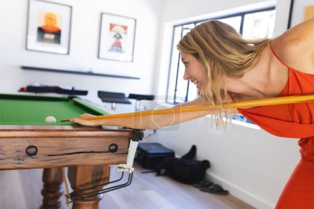 Young Caucasian woman plays pool in a bright room with art on the walls. Dressed in an orange dress, she focuses intently on her shot, leaning over the pool table.