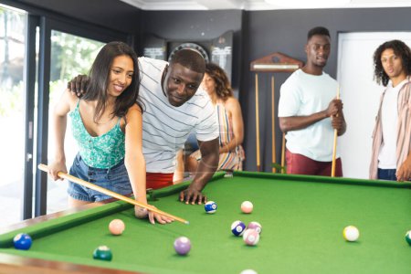 Biracial woman and African American man play pool as another African American man watches. Casual clothing and relaxed atmosphere suggest a leisurely social gathering.