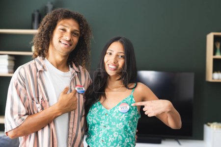 Biracial couple proudly displaying "I Voted" stickers, standing in a home setting. Young man with curly hair and woman in a green floral top express civic engagement with bright smiles.