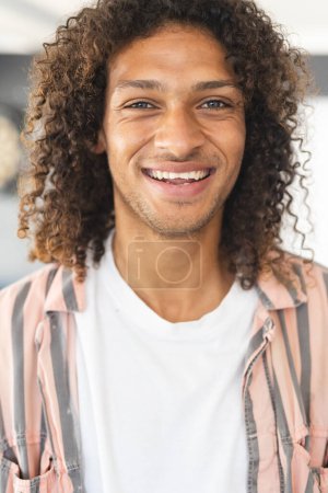 Young biracial man with curly brown hair smiles warmly. He wears a white t-shirt with a striped pink overshirt, radiating a casual, friendly vibe.